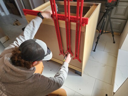 With the Cabinet Plumbing Jig leveled and centered on the cabinet layout, a worker aligns the arms with the pipes (right). After tightening the arms in place, he moves the jig to the back of the cabinet to mark out the pipe locations (above).