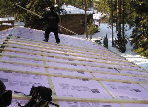 Crew members slide long 2x4 spacers into the C-shaped perforated metal vent fastened to the bottom edge of the roof. They run the spacers long, then cut them in place at the ridge.